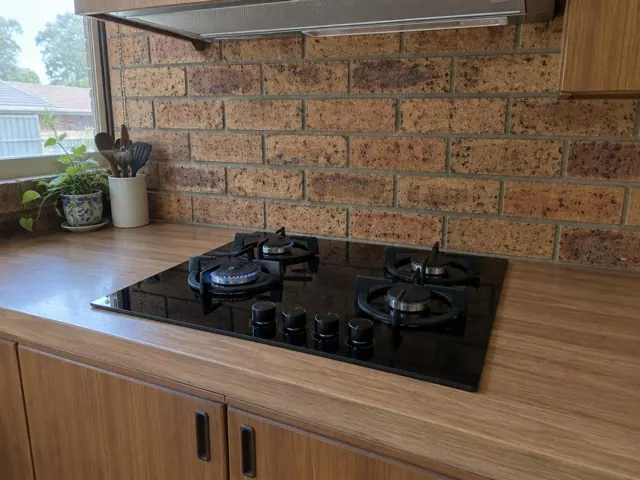 Black gas cooktop on a wood-look bench with brick backsplash and window light in Ashtonfield