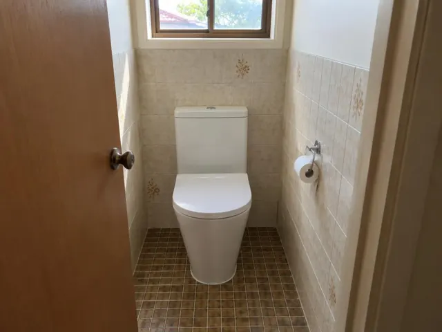 White toilet in a small tiled bathroom with a window and brown floor tiles in Metford